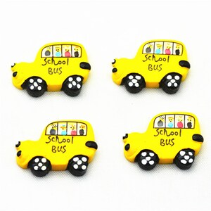 Op de afbeelding: Vier gele decoratieve magneten in de vorm van een schoolbus. Elke bus heeft zwarte wielen met witte accenten en een raam met kleurrijke figuren. De woorden "School Bus" staan in het zwart op elke bus.