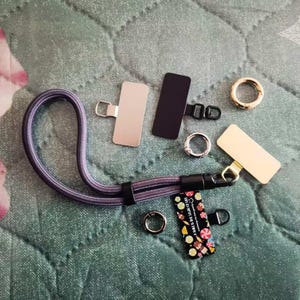 Pode incluir: Um conjunto de ferragens metálicas sortidas para artesanato, incluindo um retângulo preto, um retângulo prateado, um retângulo dourado, um clipe preto e prateado, um anel prateado, um gancho giratório prateado e um cordão preto e prateado com uma alça trançada roxa e cinza. O cordão tem uma etiqueta colorida com o texto "A LITTLE BIT OF MAGIC" impresso nela.