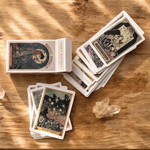 May include: A deck of Magic Rituals tarot cards with a white box featuring a circular illustration of a figure. The cards display intricate, colorful artwork with celestial and floral motifs, accompanied by two clear quartz crystals.