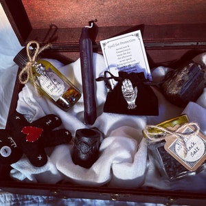 May include: A wooden box filled with various witchy items, including a black candle, a small bottle of oil, a black crystal, a black salt jar, a black voodoo doll, and a small pouch with a silver hand charm. The box is lined with white fabric and the items are arranged neatly.