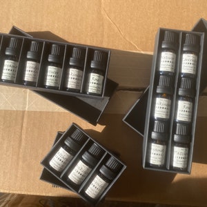 May include: Six sets of small glass bottles with black labels and white text. The bottles are arranged in rows in black boxes. The labels on the bottles read "Patchouli", "Orange", "Lavender", "Frankincense", "Rose", "Vetiver", "Juniper", "Tea Tree", "Peppermint", and "Patchouli".