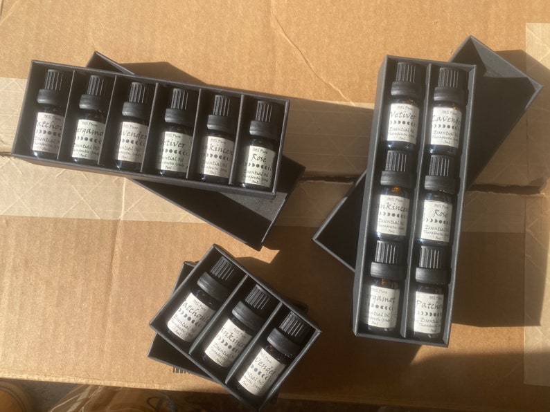 May include: Six sets of small glass bottles with black labels and white text. The bottles are arranged in rows in black boxes. The labels on the bottles read "Patchouli", "Orange", "Lavender", "Frankincense", "Rose", "Vetiver", "Juniper", "Tea Tree", "Peppermint", and "Patchouli".