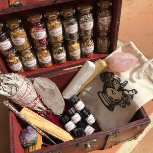 Puede incluir: Un cofre de madera roja lleno de artículos espirituales. Contiene pequeños frascos de vidrio con hierbas, un manojo de salvia, cristales, botellas de aceites esenciales, un cuarzo rosa, una varita y una pequeña bolsa con un dibujo de una mano.