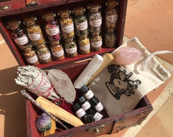 May include: A wooden chest filled with various spiritual items. It contains small glass jars with herbs, a bundle of sage, crystals, essential oil bottles, a rose quartz, a wand, and a small bag with a hand graphic.