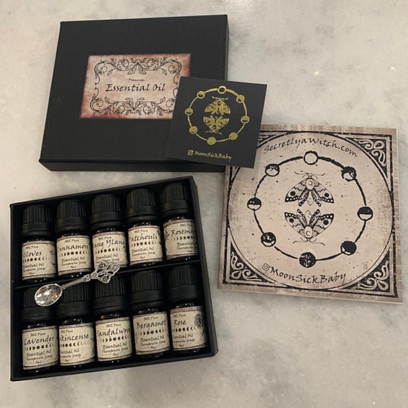 May include: A set of 10 small, black glass bottles with gold labels containing essential oils. The bottles are arranged in a black box with a gold label that reads "Premium Essential Oil". The box is sitting on a white surface. The image also includes a small, gold spoon and a card with a black and white illustration of moths and moons.