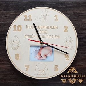 May include: A round wooden clock with a light-colored face. The clock features a photo of a baby, numbers, and the text "DZIĘKU KOCHANYM CIOCIOM A OPIEKĘ PIOTRUŚ 24.0 2019 1770g I 47cm". The clock has black and red hands and baby illustrations.