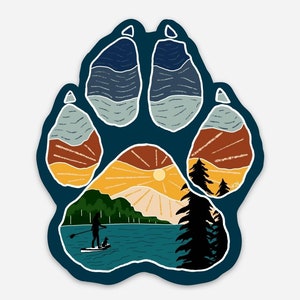 Puede incluir: Una huella de pata azul con una escena de puesta de sol en su interior. La escena incluye un lago, monta&ntilde;as, &aacute;rboles y una persona haciendo paddleboard con un perro.