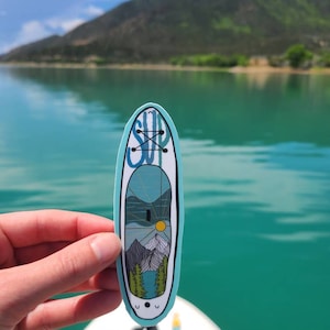 Puede incluir: Una pegatina azul y blanca con una escena de montaña y la palabra "SUP" en azul. La pegatina se está sosteniendo frente a un lago con montañas en el fondo.