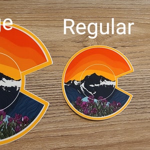 May include: Three Colorado mountain sunset stickers in different sizes: large, regular, and mini. The stickers feature a stylized mountain range with a sunset sky and wildflowers.