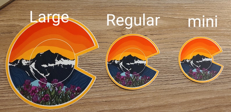 May include: Three Colorado mountain sunset stickers in different sizes: large, regular, and mini. The stickers feature a stylized mountain range with a sunset sky and wildflowers.