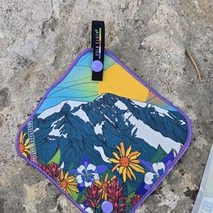 May include: A diamond-shaped cloth with a mountain and floral design. The cloth features a purple border and a black strap with the text "KULA CLOTH". The design includes a mountain range, a yellow sun, and colorful flowers.
