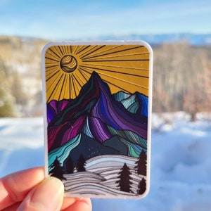 May include: A sticker featuring a colorful, line art illustration of a mountain range with a sun in the background. The mountains are purple, blue, and green, and the sun is yellow. The sticker is on a white background.