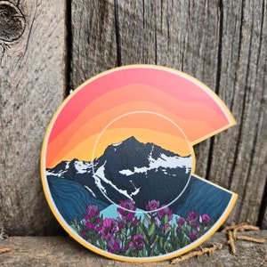 May include: A colorful sticker featuring a stylized outline of the state of Colorado with a mountain range, sunset, and wildflowers. The sticker is on a wooden surface.
