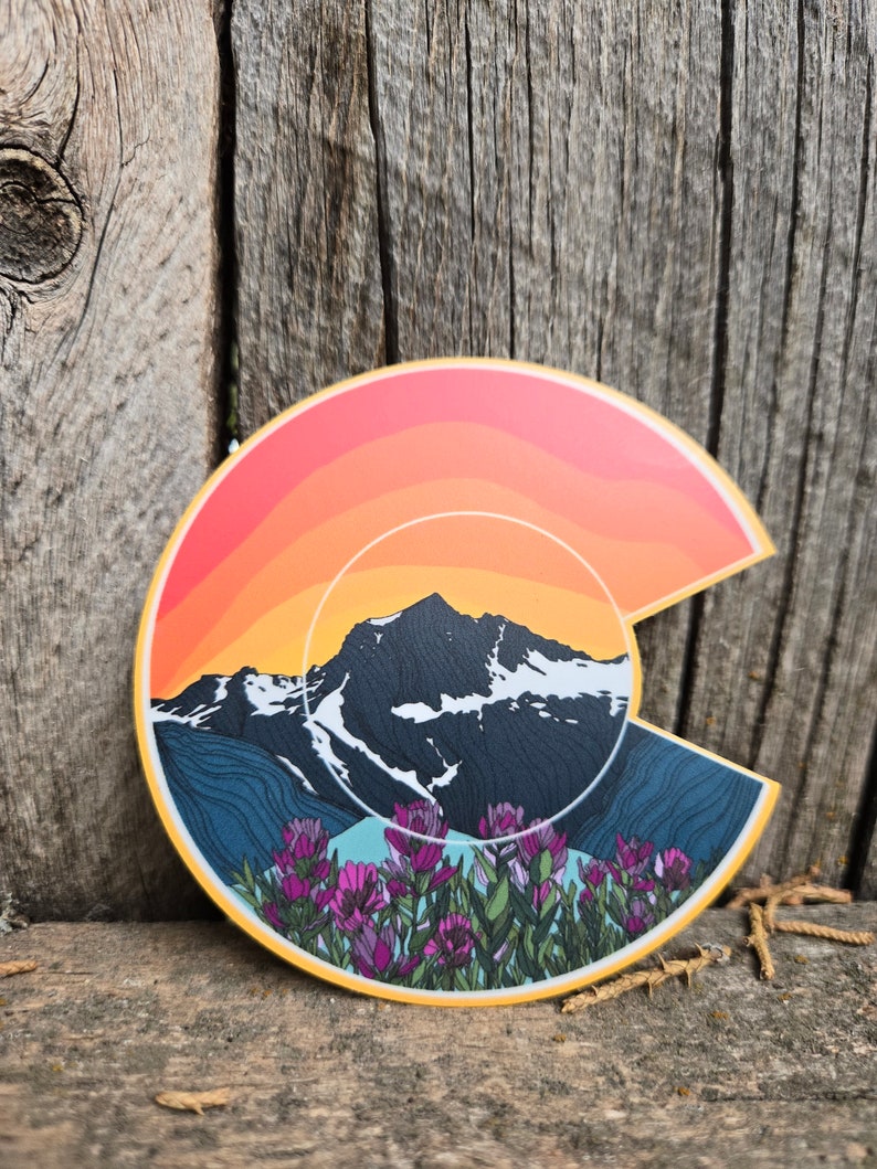 May include: A colorful sticker featuring a stylized outline of the state of Colorado with a mountain range, sunset, and wildflowers. The sticker is on a wooden surface.