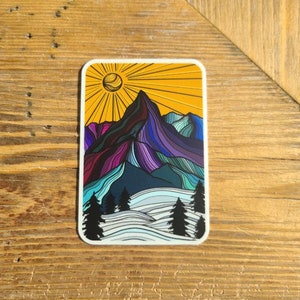 May include: A colorful sticker featuring a stylized mountain range with a sun shining above. The mountain is purple, blue, and green, with a yellow sun and white snow at the base.