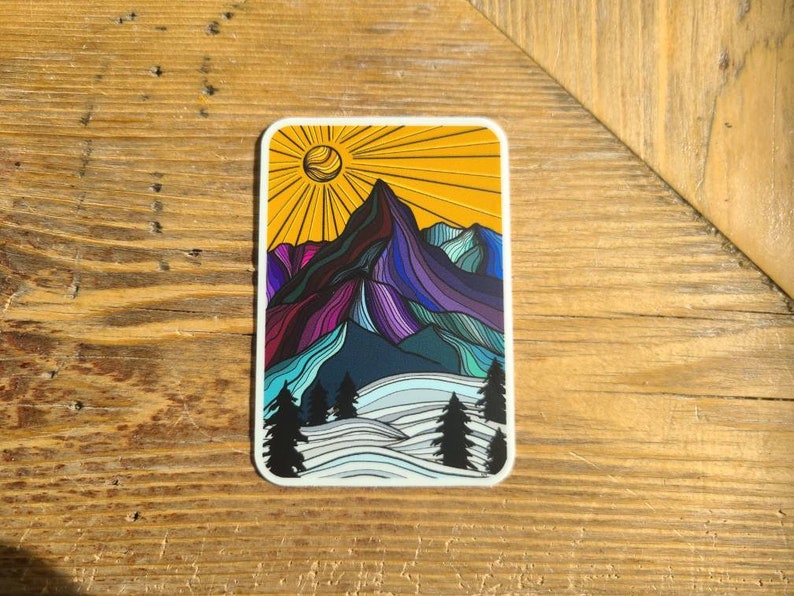 May include: A colorful sticker featuring a stylized mountain range with a sun shining above. The mountain is purple, blue, and green, with a yellow sun and white snow at the base.