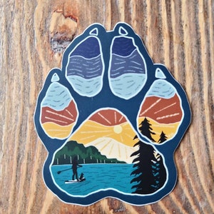 Puede incluir: Una pegatina azul en forma de huella de pata con una escena de puesta de sol en su interior. La escena presenta un lago, monta&ntilde;as, un sol y una persona haciendo paddleboard con un perro.