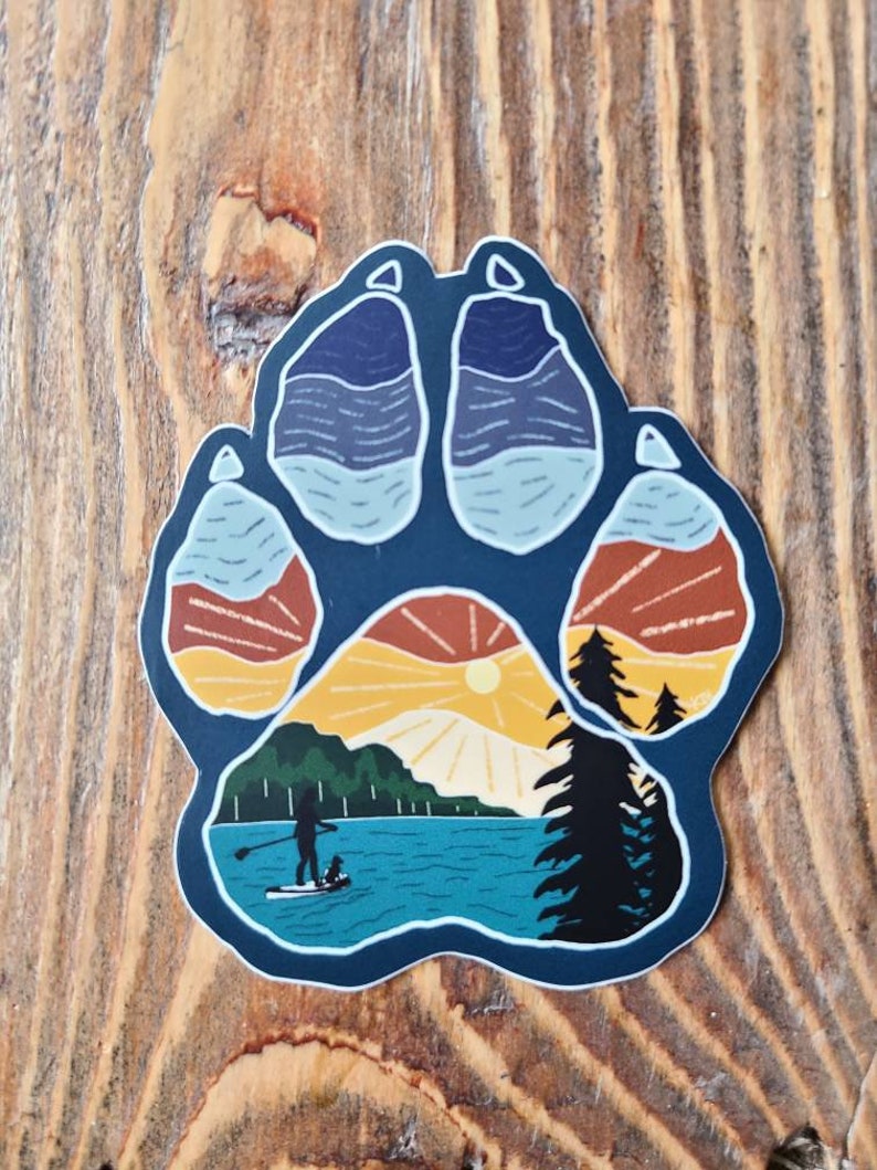 Puede incluir: Una pegatina azul en forma de huella de pata con una escena de puesta de sol en su interior. La escena presenta un lago, monta&ntilde;as, un sol y una persona haciendo paddleboard con un perro.