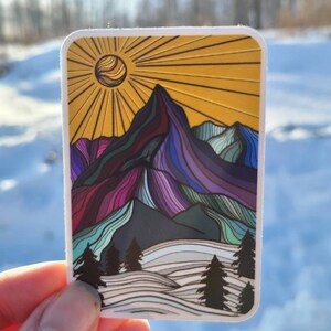 May include: A sticker with a colorful mountain landscape design. The mountains are outlined in black and filled with shades of purple, blue, green, and pink. The sun is yellow with orange rays. The background is white with a blue sky.