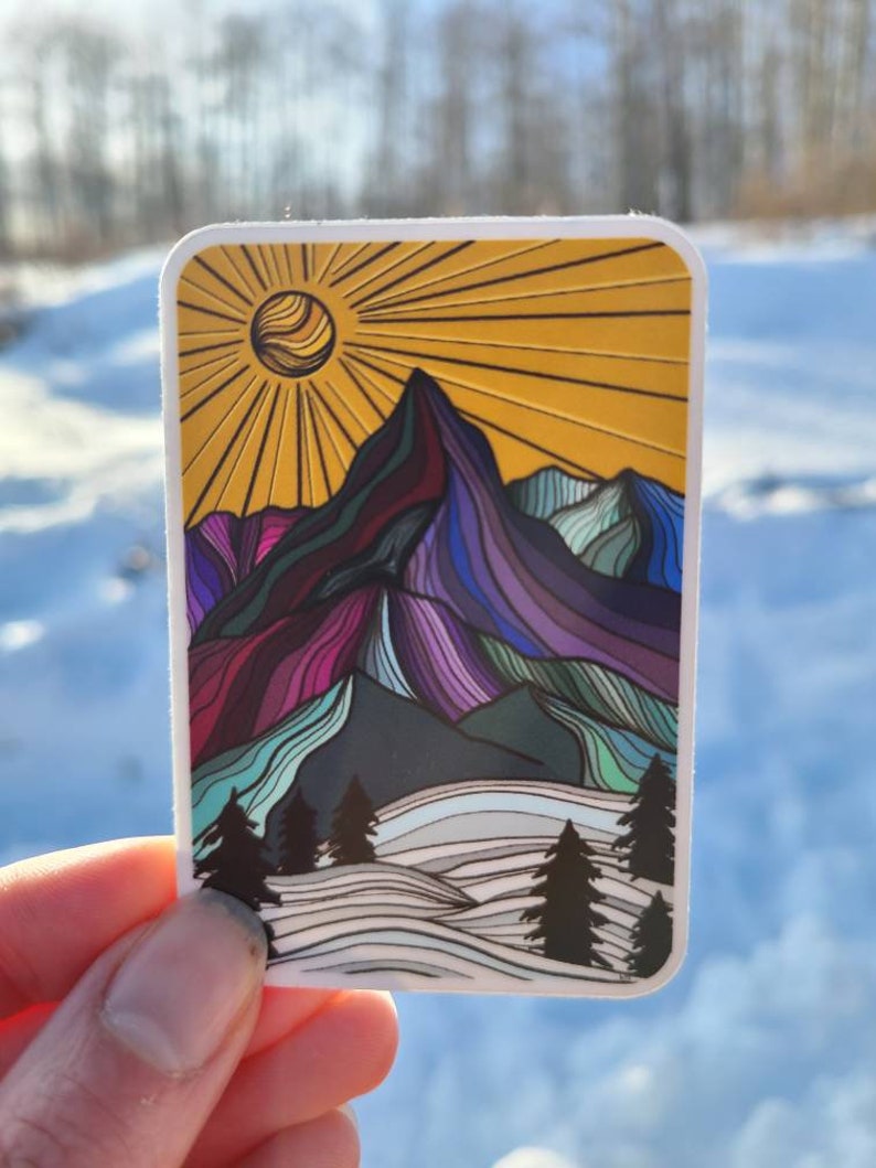 May include: A sticker with a colorful mountain landscape design. The mountains are outlined in black and filled with shades of purple, blue, green, and pink. The sun is yellow with orange rays. The background is white with a blue sky.
