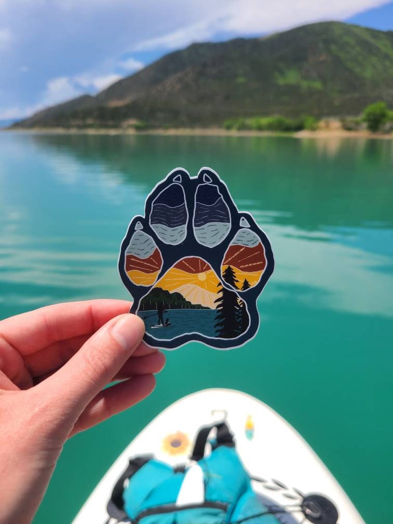 Puede incluir: Una pegatina de huella de pata azul con una escena de puesta de sol en su interior. La escena incluye un lago, monta&ntilde;as y una persona haciendo paddleboard.