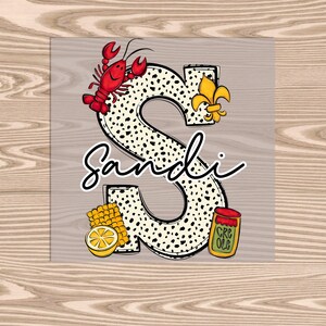 May include: A white and black polka dot letter S with a red crawfish, a yellow lemon, a yellow corn cob, and a red and yellow jar of Creole seasoning. The letter S is decorated with a gold fleur-de-lis. The word "Sandi" is written in black cursive below the letter.