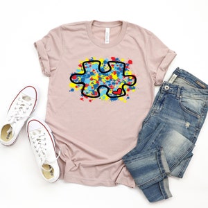 Op de afbeelding: Een lichtroze T-shirt met een zwarte omtrek van een puzzelstuk dat is gevuld met kleurrijke verfspatten. Het puzzelstuk heeft een klein rood hartje in het midden. Het T-shirt ligt op een wit oppervlak met een paar witte sneakers en een blauwe jeans.