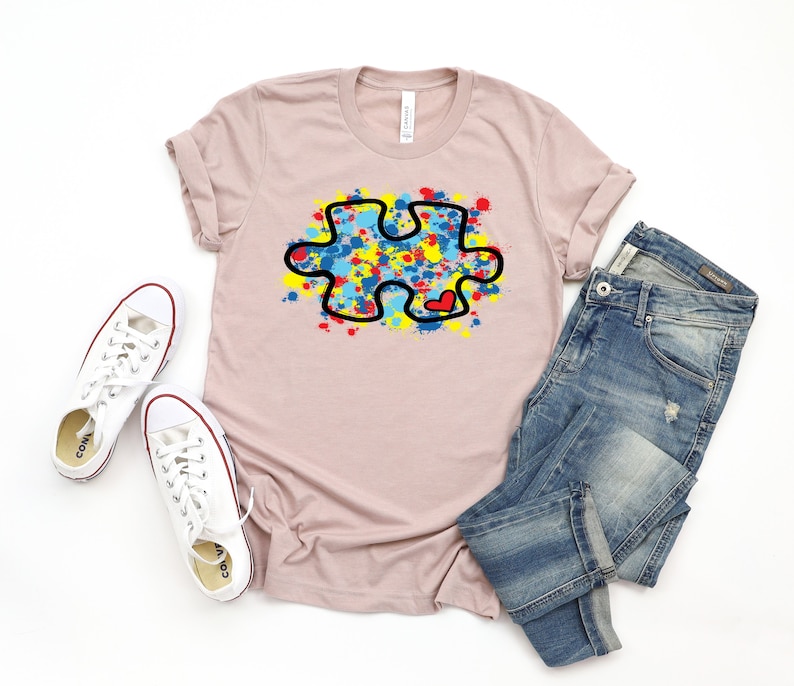 Op de afbeelding: Een lichtroze T-shirt met een zwarte omtrek van een puzzelstuk dat is gevuld met kleurrijke verfspatten. Het puzzelstuk heeft een klein rood hartje in het midden. Het T-shirt ligt op een wit oppervlak met een paar witte sneakers en een blauwe jeans.