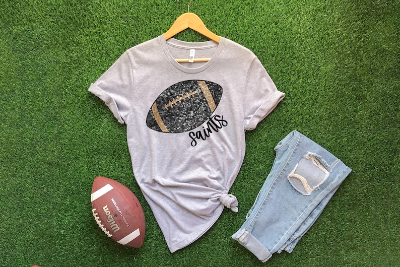 May include: A light gray t-shirt with a football graphic and the word "Saints" in black script. A brown and white football and a pair of blue jeans are also in the image. The shirt is displayed on a green surface.