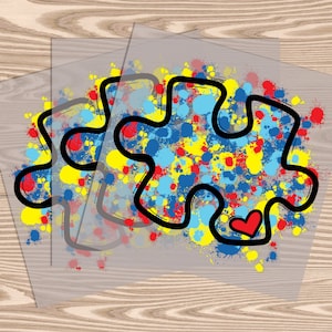 Op de afbeelding: Twee transparante vinyl stickers met een zwarte omtrek van een puzzelstuk. Het puzzelstuk is gevuld met kleurrijke verfspatten in tinten blauw, geel en rood. Een klein rood hartje bevindt zich in het puzzelstuk.