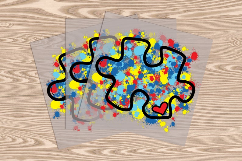 Op de afbeelding: Twee transparante vinyl stickers met een zwarte omtrek van een puzzelstuk. Het puzzelstuk is gevuld met kleurrijke verfspatten in tinten blauw, geel en rood. Een klein rood hartje bevindt zich in het puzzelstuk.
