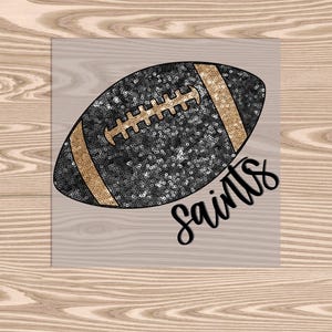 May include: A black and gold football with the word "Saints" in a script font. The football is covered in sequins and has gold stripes and laces. The image is on a light wood grain background.