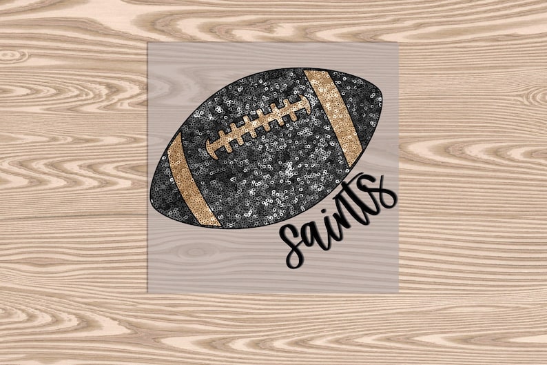 May include: A black and gold football with the word "Saints" in a script font. The football is covered in sequins and has gold stripes and laces. The image is on a light wood grain background.
