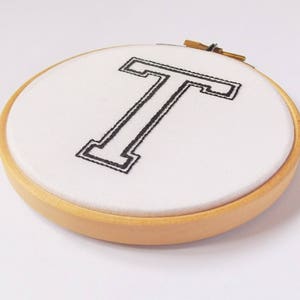 May include: A white fabric hoop with a black embroidered letter T in a varsity style font. The hoop is made of wood and has a natural finish.