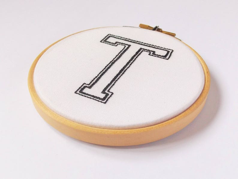 May include: A white fabric hoop with a black embroidered letter T in a varsity style font. The hoop is made of wood and has a natural finish.