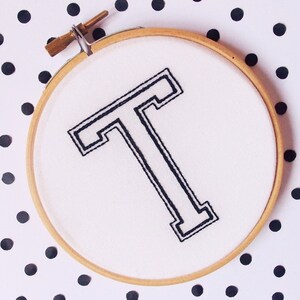 May include: A white embroidery hoop with a black embroidered capital letter "T" in a bold, outlined font. The hoop is on a white background with black polka dots.
