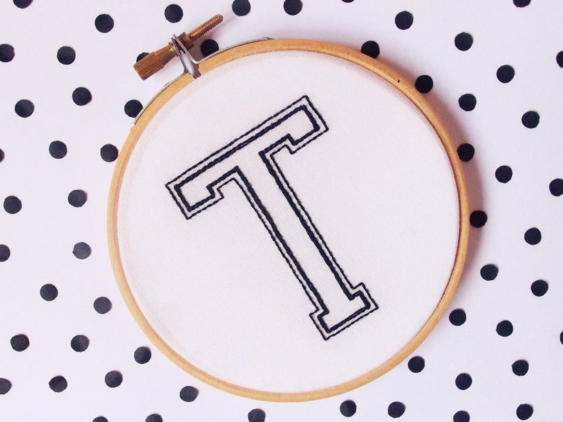 May include: A white embroidery hoop with a black embroidered capital letter "T" in a bold, outlined font. The hoop is on a white background with black polka dots.