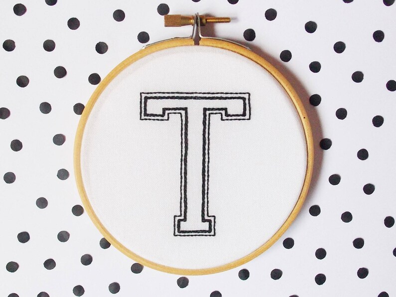 May include: A white embroidery hoop with a black embroidered letter "T" on a white background. The hoop is on a white background with black polka dots.