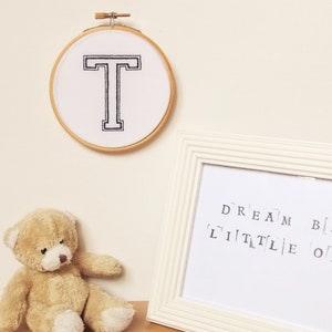 May include: A white embroidery hoop with a black embroidered letter T. A white frame with the text "DREAM BIG LITTLE ONE" is on a white surface with a brown teddy bear.