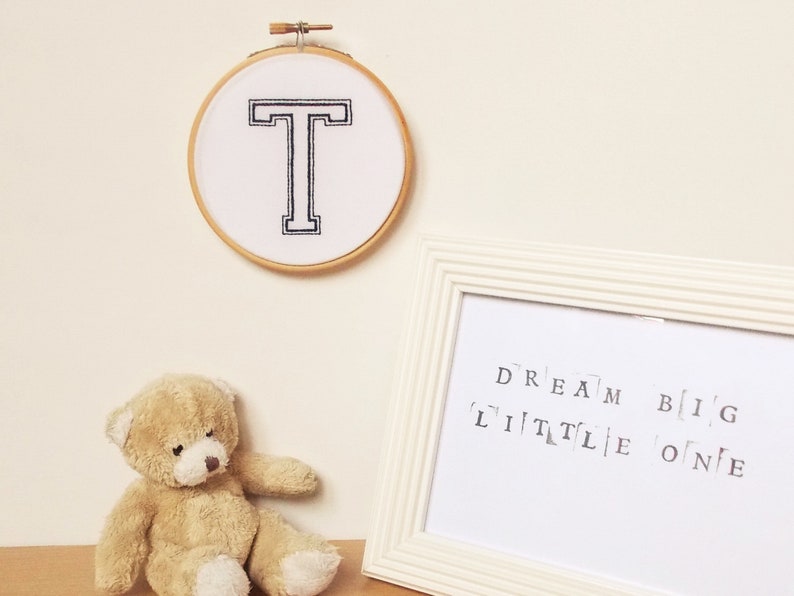 May include: A white embroidery hoop with a black embroidered letter T. A white frame with the text "DREAM BIG LITTLE ONE" is on a white surface with a brown teddy bear.