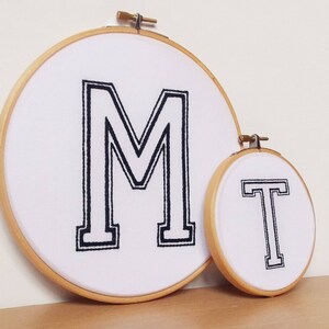 May include: Two embroidery hoops with white fabric and black embroidery. The larger hoop has the letter "M" embroidered in a bold, blocky font. The smaller hoop has the letter "T" embroidered in the same font.