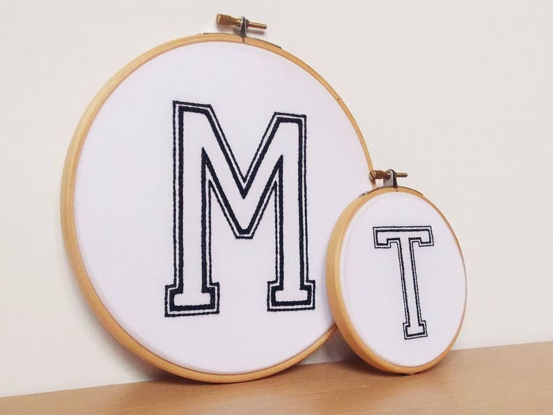 May include: Two embroidery hoops with white fabric and black embroidery. The larger hoop has the letter "M" embroidered in a bold, blocky font. The smaller hoop has the letter "T" embroidered in the same font.