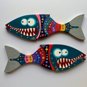 May include: Two whimsical, hand-painted wooden fish decorations. Each fish has a teal body, large white eyes, and a wide, toothy grin. The fins are decorated with colorful patterns, and the tails are gray with horizontal lines.