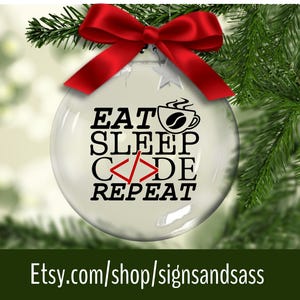 May include: A clear glass Christmas ornament with the text "EAT SLEEP CODE REPEAT" in black, a coffee cup graphic, and a red bow. The ornament is hanging from a green Christmas tree branch.