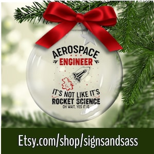 May include: A clear glass Christmas ornament with a red bow and green pine branches. The ornament has the text "AEROSPACE ENGINEER" and "IT'S NOT LIKE IT'S ROCKET SCIENCE OH WAIT, YES IT IS" with a rocket ship graphic. The Etsy shop URL is visible.