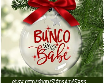 May include: A clear glass Christmas ornament with the words "Bunco Babe" in red script, along with dice illustrations. The ornament is adorned with a red bow and star-shaped accents, and is hanging on a green Christmas tree branch.