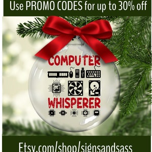 May include: A clear glass Christmas ornament with the words "COMPUTER WHISPERER" in red. The ornament features black icons of computer components and is topped with a red bow. The ornament hangs from a green pine branch.