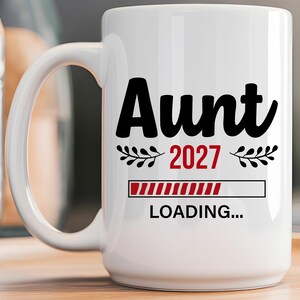 May include: A white ceramic mug featuring the black text Aunt, the red text 2027, and a red loading bar graphic with the black text LOADING... underneath. The design includes small black leaf accents on either side of the year 2027.