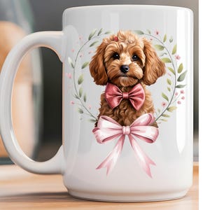 May include: A white ceramic mug featuring a graphic of a brown curly-haired puppy wearing a pink bow tie. The puppy is centered within a circular wreath of green leaves and small pink flowers, with a large pink ribbon tied at the bottom of the design.