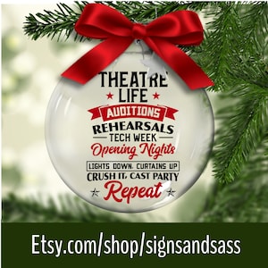 May include: A clear glass Christmas ornament with a red bow, featuring the words "THEATRE LIFE" and other theater-related terms like "Auditions" and "Opening Nights." The ornament is hanging among green pine needles. The Etsy shop URL is visible at the bottom.